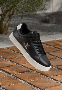 Sneaker in pelle nera di Calvin Klein con suola e linguetta del tallone bianche, lacci neri, esposto sopra piastrelle quadrate di legno all'aperto.