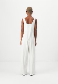 Combinaison blanche à jambes larges avec un dos carré et un panneau en tissu texturé. Caractérisée par de fines bretelles et un drapé fluide.