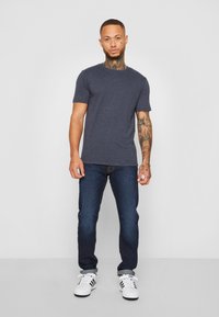Marineblaues T-Shirt mit kurzen Ärmeln, aus mittelschwerem Stoff, kombiniert mit dunkelblauen Jeans. Weiße Sneaker haben schwarze Akzente und Streifen.