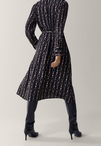 Musta puoliksi läpinäkyvä mekko pitkillä hihoilla, jossa on kultainen ja vaaleanpunainen ketju kuvio. Vyötärölle vyötetty ja virtaava siluetti.