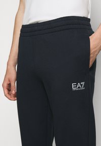 Pantalons de survêtement bleu marine avec une taille côtelée, des poches latérales et un petit logo EA7 blanc sur la cuisse. Le matériau semble doux et lisse.
