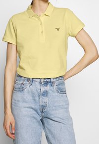 Polo shirt jaune pâle avec un col et trois boutons. Présente un petit logo noir "GANT" sur la poitrine. Associé à un jean en denim bleu clair.