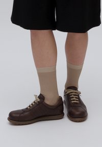 Person wearing beige mid-calf socks and dark brown leather casual shoes with light brown laces, standing against a white background.
