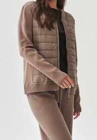 Giacca a vento marrone in tessuto di maglia. Presenta una zip frontale, polsini e orlo a coste. Texture liscia con design a pannelli trapuntati.