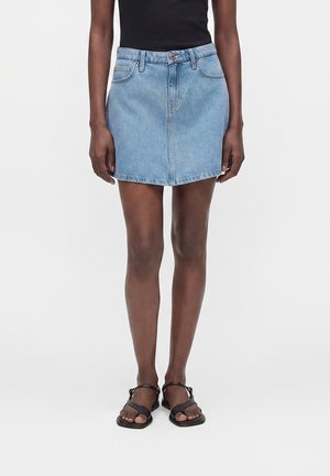 Ansicht von der Taille abwärts einer Person, die einen hellblauen Jeansrock, ein schwarzes ärmelloses Top und schwarze flache Sandalen mit Knöchelriemen trägt.