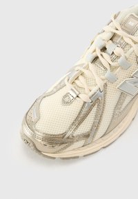 Cream-colored sneaker with metallic silver accents, mesh material, textured surface, lace-up design, and logo detail on the side.