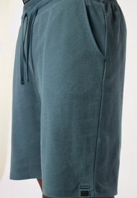 Grijs-blauwe textuurshorts met een elastische tailleband, trekkoord, zijzakken en een klein logo-label nabij de zoom.