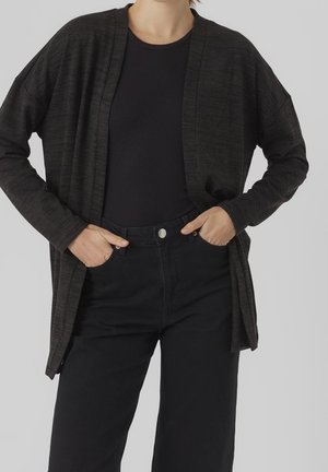 Person iført en mørk cardigan med åben front over en sort top og sorte bukser med høj talje med hænderne i lommerne mod en grå baggrund.