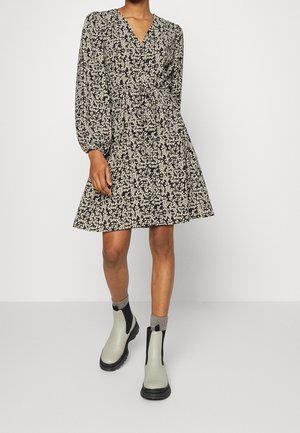 Femme portant une robe à manches longues noire et blanche à motif floral avec un col en V, assortie à des chaussettes grises et des bottines grises claires.