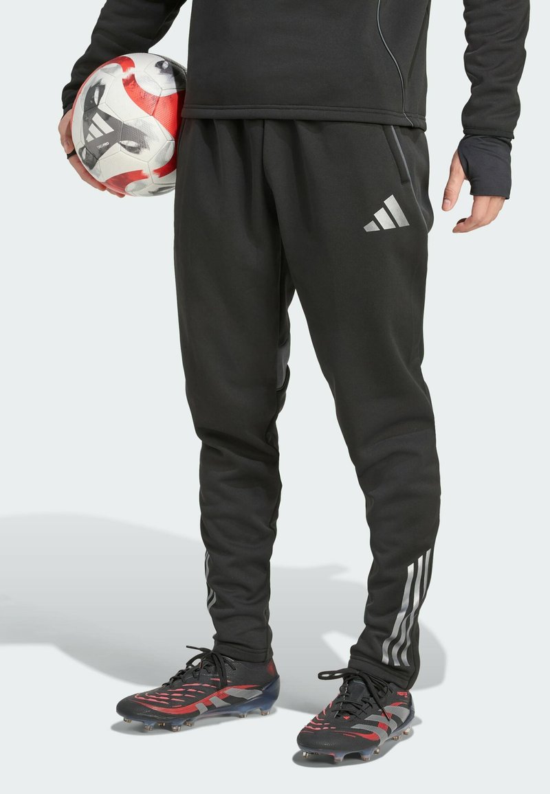 Schwarze Adidas-Sweatpants mit taillierter Passform, die an den Seiten mit drei silbernen Streifen verziert sind, kombiniert mit roten und schwarzen Fußballschuhen.