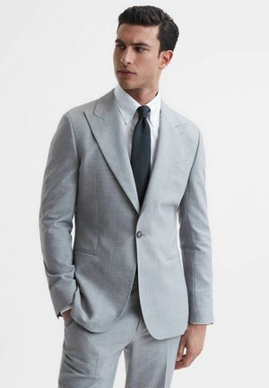 Uomo con abito grigio chiaro, camicia bianca e cravatta nera, in piedi con una mano in tasca, che guarda verso sinistra.