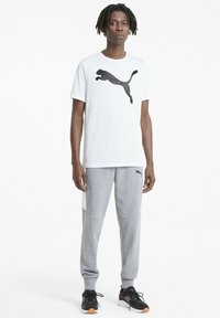 Balta īsām piedurknēm T-kreklu ar melnu Puma logo, apvienojot ar pelēkiem sporta biksēm ar melniem un baltajiem akcentiem un melnām sporta kedām.