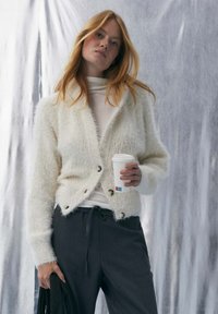 Weißer, flauschiger Cardigan mit V-Ausschnitt und Knopfverschluss. Wird über einem Rollkragenpullover getragen und mit dunklen, locker sitzenden Hosen kombiniert. Hält eine Kaffeetasse.