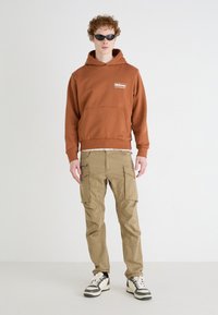 Brązowa bluza z kapturem z przednią kieszenią i logo, zestawiona z khaki spodniami cargo z bocznymi kieszeniami. Model nosi czarne okulary przeciwsłoneczne i trampki.