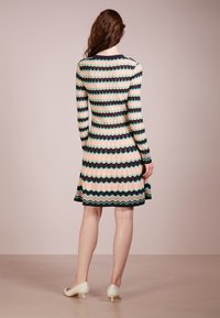 Ribbad klänning med zigzagmönster i marinblått, rosa och grönt. Långa ärmar, figursydd siluett, utställd fåll och rund halsringning. Bärs med ljusa klackar.