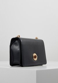 Černá kožená kabelka s texturovaným povrchem, zlatými kovovými detaily, kulatým zapínáním a pevně tvarovanou konstrukcí.