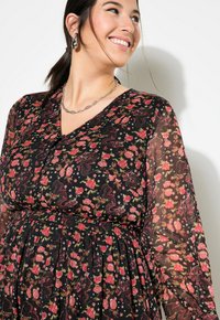 Usměvavá žena s tmavými vlasy v culíku na sobě má černé květinové šaty s růžovými květy a stříbrný řetízkový náhrdelník, hledí do strany.