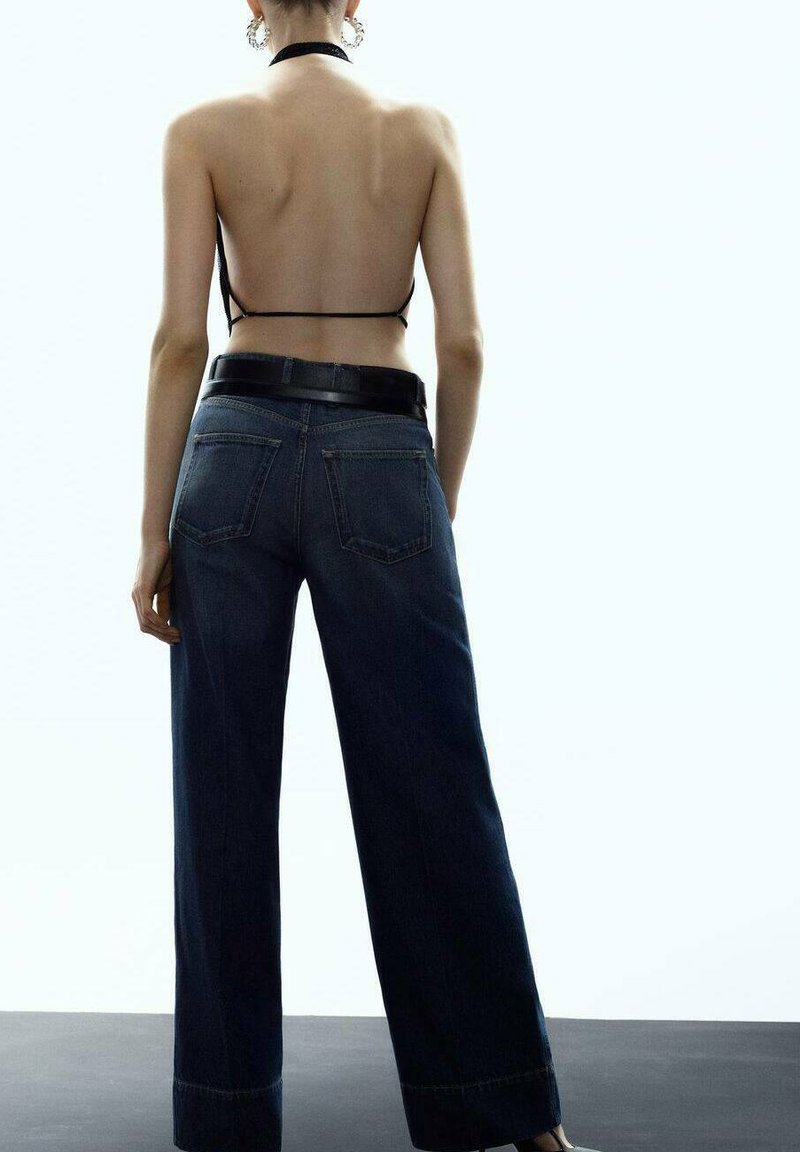 Tylny widok modelki w ciemnoniebieskich spodniach o szerokich nogawkach, z czarnym skórzanym paskiem i cienkim czarnym topem na szyję. Widoczna gładka tekstura materiału.