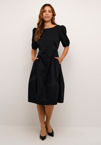 Robe en coton noir avec un col rond, des manches bouffantes et des poches latérales ; présente une jupe évasée et une texture lisse.