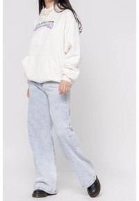 Krémová mikina s kapucí, přední kapsou a šňůrkou, spárovaná se světle modrými širokými džíny a černými dřeváky s hnědou podrážkou.