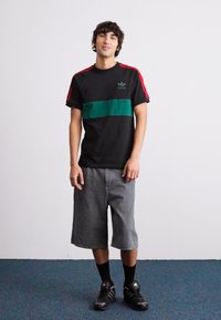 Svart kortärmad skjorta med gröna horisontella ränder och röda detaljer; grå knälånga shorts; svarta sneakers och strumpor; neutral bakgrund.