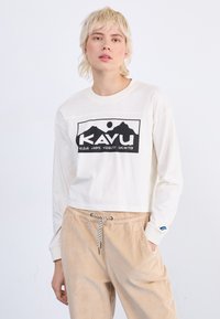 Weißes Langarmhemd mit schwarzem Berggrafik und "KAVU"-Text. Kombiniert mit beigen Cordhose mit Kordelzug.