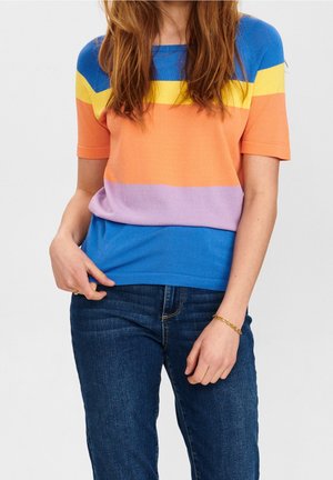 Woman wearing a short-sleeve sweater with blue, yellow, orange, and purple stripes and dark blue jeans, with a gold bracelet on left wrist.