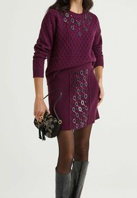 Pull en maille violet avec motif texturé, associé à une jupe assortie ornée d'accents circulaires décoratifs. Sac à main noir perlé et bottes montantes.