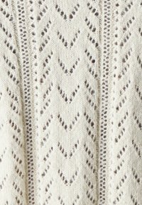 Cremfarbener Strickstoff mit Zickzackmuster und offenen Details, die ein strukturiertes Aussehen erzeugen. Weiches, leichtes Material.