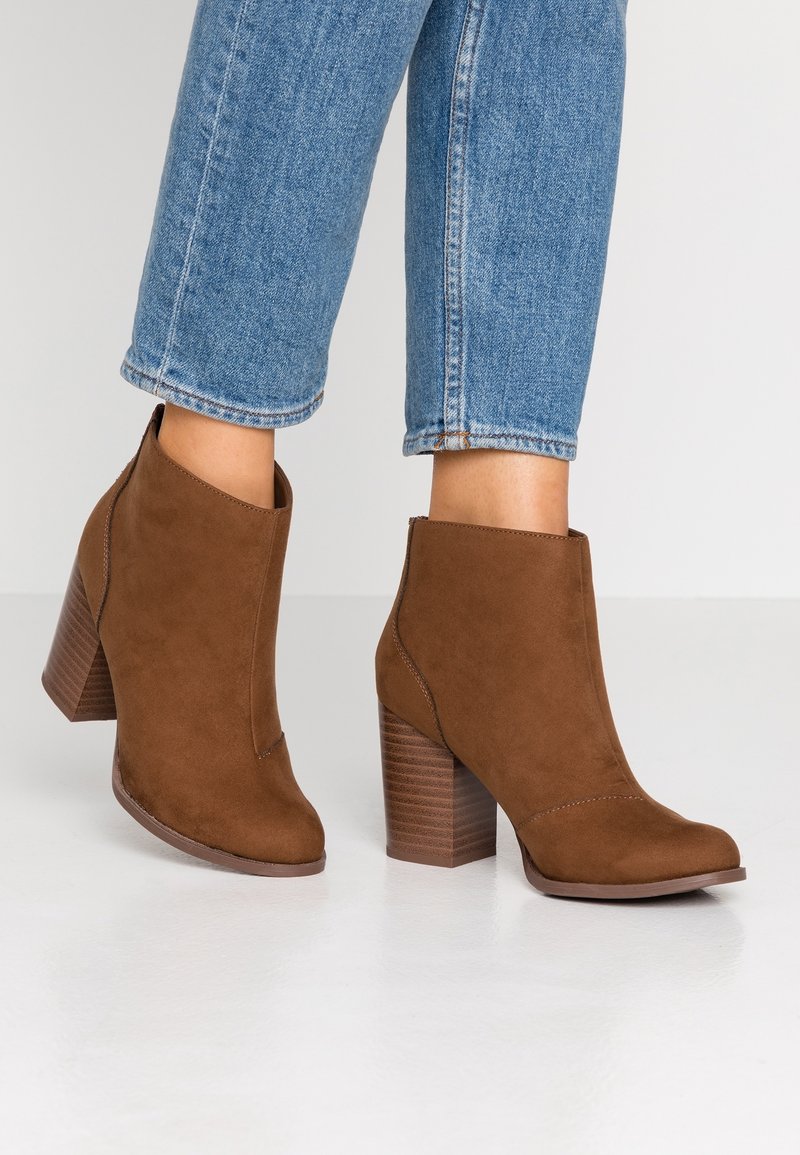 Mujer con botas de ante marrón hasta el tobillo con tacones cuadrados y jeans cortos azules sobre un suelo blanco.