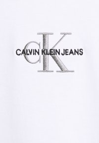 Camiseta blanca de algodón con el logo de CALVIN KLEIN JEANS en negro y un bordado metálico plateado de "CK", textura suave y diseño de cuello redondo.