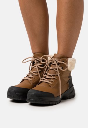 Bottes brunes imperméables avec semelle en caoutchouc noire, design à lacer et doublure en fausse fourrure au niveau du col. Matériau supérieur imperméable texturé.