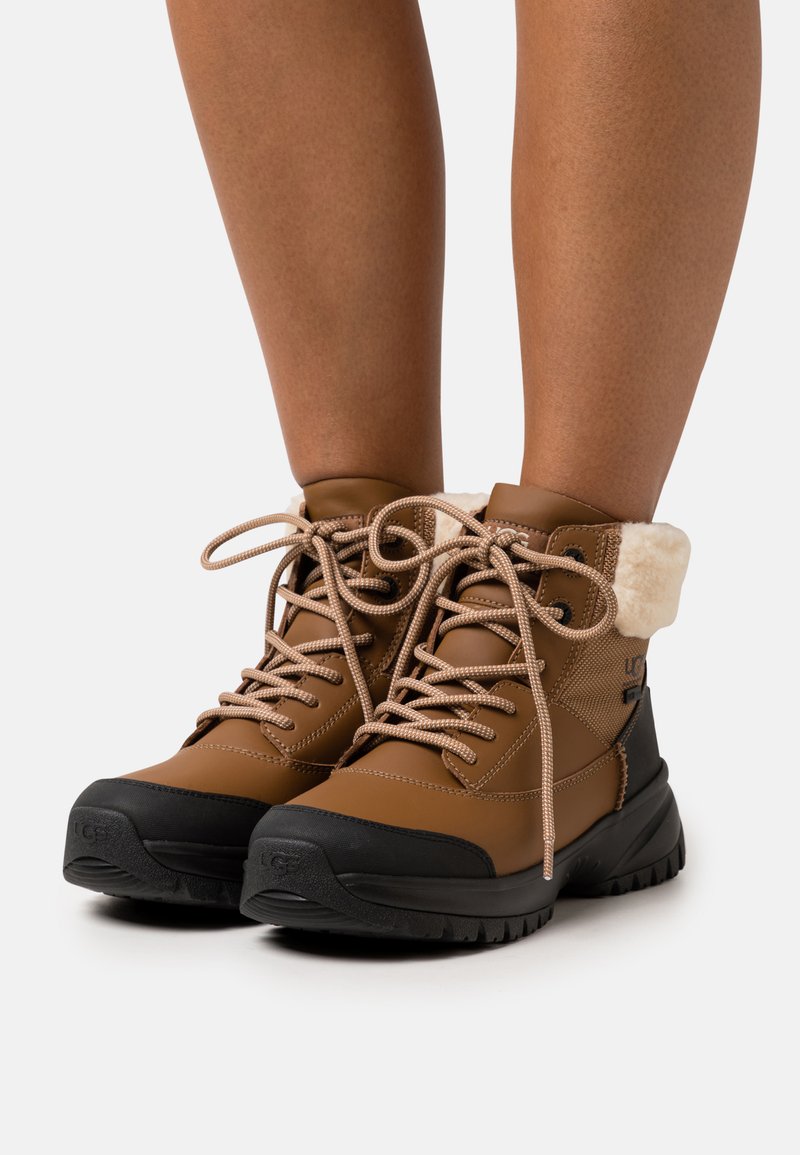 Botas marrones impermeables con suela negra de goma, diseño con cordones y forro de piel sintética en el collar. Material superior impermeable texturizado.