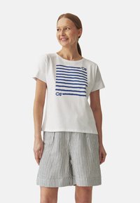 T-shirt en coton blanc avec des rayures horizontales bleues et un design de nœuds, associé à un short plissé rayé noir et blanc.