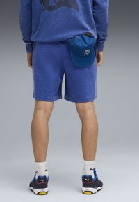 Felpa blu con motivo scuro, pantaloni corti blu e cappellino da baseball blu in tasca. Le sneaker presentano un design nero e blu con accenti bianchi.