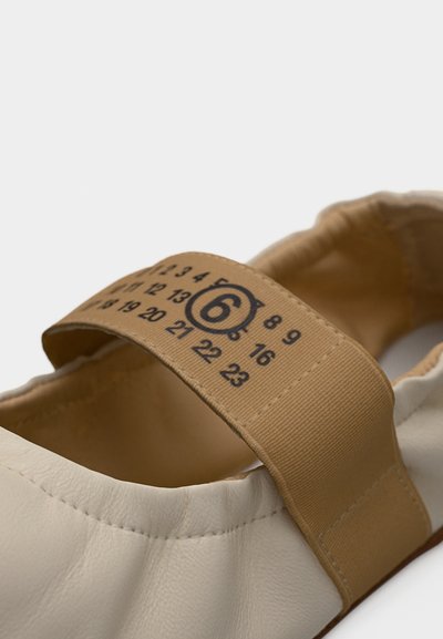 Κοντινό πλάνο σε απαλό παπούτσι μπεζ με ελαστικό λουράκι που φέρει μαύρους τυπωμένους αριθμούς και κυκλωμένο τον αριθμό 6 στο λουράκι.
