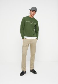 Green sweatshirt with white and red logo, beige pants, and black shoes. The outfit is completed with a gray cap featuring a back strap.