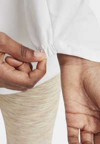 Camisa blanca de manga larga con un acabado texturizado, que presenta puños elásticos. Se tira de la tela cerca del dobladillo de la manga, revelando leggings ajustados.