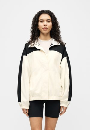 Woman with curly hair wearing a cream and black zip-up jacket and black cycling shorts, standing against a plain white background.