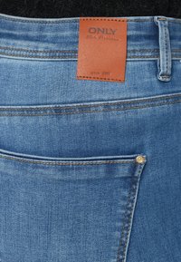 Kék farmernadrág barna bőr folttal, amelyen az "ONLY Blue Denim" felirat található. Két tűs varrás jellemzi, valamint egy kis arany gomb a hátsó zseben.