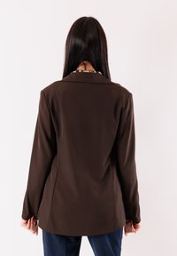 Blazer marrone con vestibilità sartoriale, maniche lunghe e colletto con rever. Presenta linee semplici e una texture liscia, senza decorazioni.