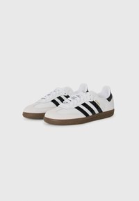 White suede sneakers featuring black stripes, a brown rubber sole, and a padded collar. The word "Samba" is printed in gold on the side.