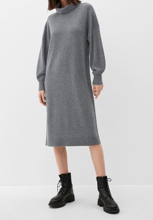 Vrouw die een grijze midi-truikleding met lange mouwen en zwarte vetercombatlaarzen draagt, staand tegen een effen witte achtergrond.