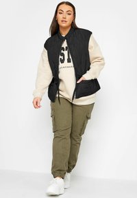 Svart vadderad väst med dragkedja framtill och sidofickor, lager på lager över en krämfärgad hoodie med tryckt text, kombinerad med olivgröna cargobyxor och vita sneakers.