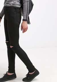 Vaqueros pitillo negros con detalles rasgados en las rodillas, hechos de denim, combinados con zapatos negros de cordones y un suéter gris texturizado.