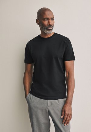 Középkorú férfi szürke szakállal, fekete pólót és szürke nadrágot viselve, egy kézzel a zsebében áll, semleges háttér előtt.