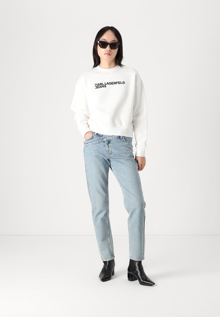 Бял суитшърт с черен надпис "KARL LAGERFELD JEANS", светлосини дънки и черни ботуши до глезена. Моделът носи черни слънчеви очила.
