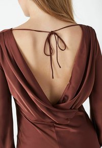 Ruda satino suknelė su žemu drapiruotu nugaros kirpimu ir ilgomis rankovėmis, turinti rišamą užsegimą kakle su minkšta, plaukiota medžiaga.