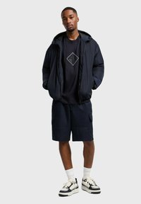 Giacca imbottita blu scuro sopra una maglietta navy con una grafica a diamante, abbinata a pantaloni corto navy e sneakers bianco e nero con suole spesse.