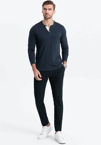 Marinblå långärmad henleytröja med vita och guldfärgade knappdetaljer, i kombination med svarta rutiga slim-fit byxor och vita sneakers.